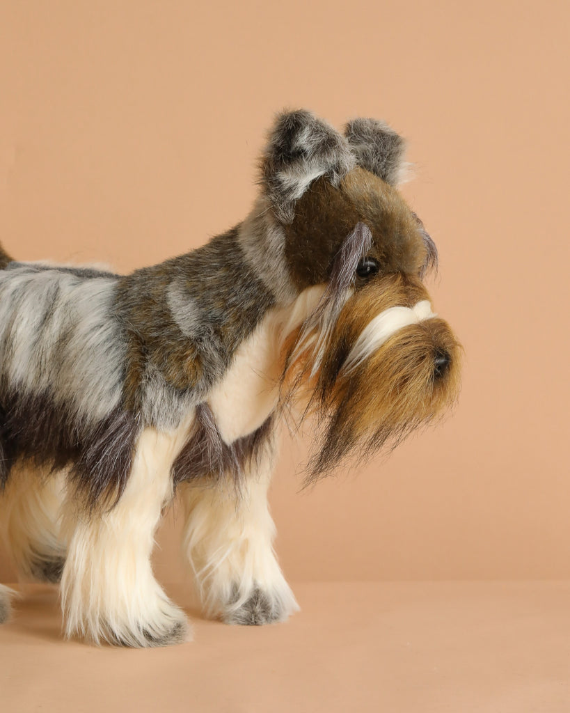 The Miniature Gray Schnauzer Dog Stuffed Animal, 18", stands on a beige surface against a beige background, featuring a long, silky gray, brown, and white coat with realistic details like a groomed beard and fluffy legs.