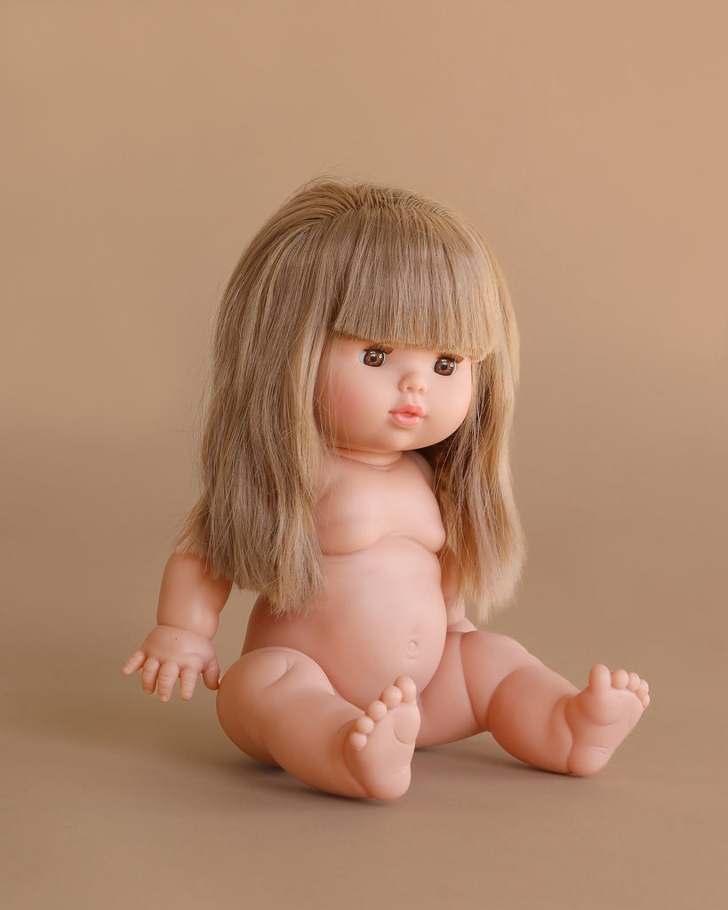 The Minikane Baby Doll (13") - Nina, featuring long, straight blonde hair and bangs, sits upright against a plain beige background. Clothing sold separately.
