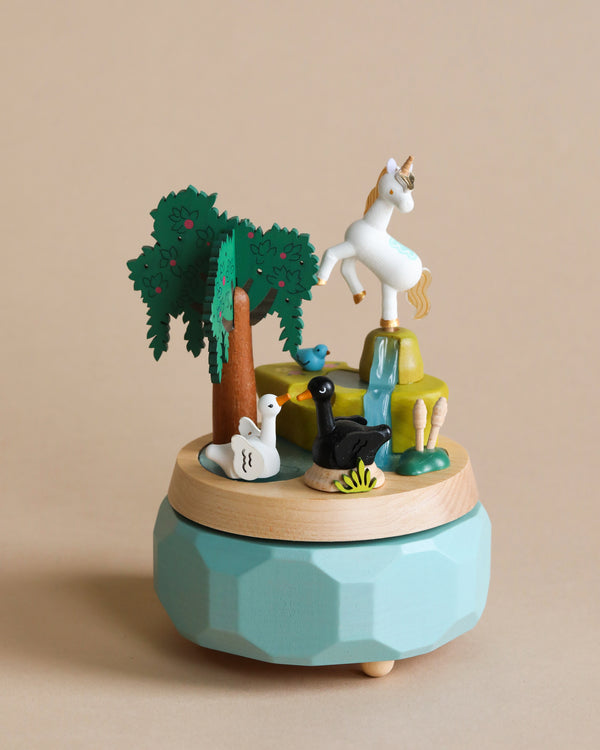 A whimsical Unicorn Music Box featuring a unicorn atop a small hill, surrounded by trees, animals, and a woman in a yoga pose, all set against a soft beige background.