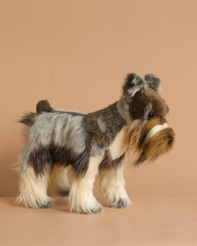 The Miniature Gray Schnauzer Dog Stuffed Animal, 18", features hand-sewn details and realistic gray, white, and brown fur, giving this plush toy a lifelike personality. Stands on a beige surface with a matching background.