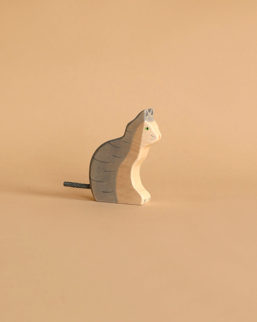 A small cat figurine from the Holztiger Handmade Wooden Animal Play Set, featuring painted gray stripes and green eyes, displays European craftsmanship on a beige surface against a matching background.