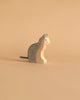 A cat figurine from the Holztiger Handmade Wooden Farm Animals Set, crafted from maple and beech wood with hand-painted gray stripes, green eyes, and a black tail, sits upright against a plain beige background.