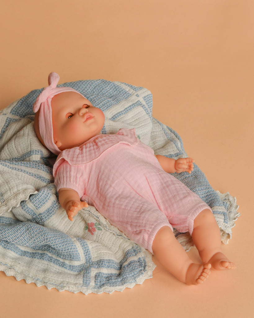 The Minikane Cuties Soft Body Doll (14") - Emilie with Clothing is dressed in a pink outfit and headband, lying on a blue and white textured blanket against a light beige background.