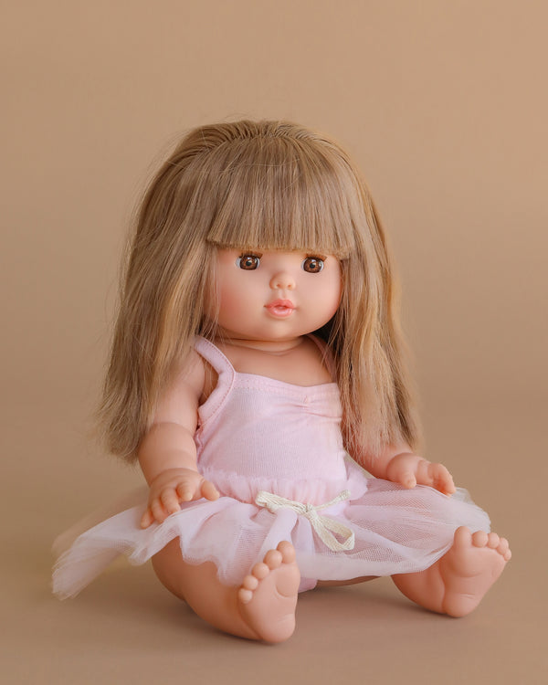 The Minikane Baby Doll (13") - Nina with Clothing features long, straight light brown hair and bangs. Made in Spain, this anatomically correct doll wears a pale pink dress with a tulle skirt and sits barefoot against a beige background.