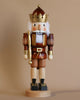 A Christian Ulbricht Nutcracker - King (Natural), designed as a soldier with a brown and gold uniform, a white beard, and a golden crown, stands on a round base against a beige background. This exquisite piece evokes the charm of the classic Christian Ulbricht Nutcrackers.