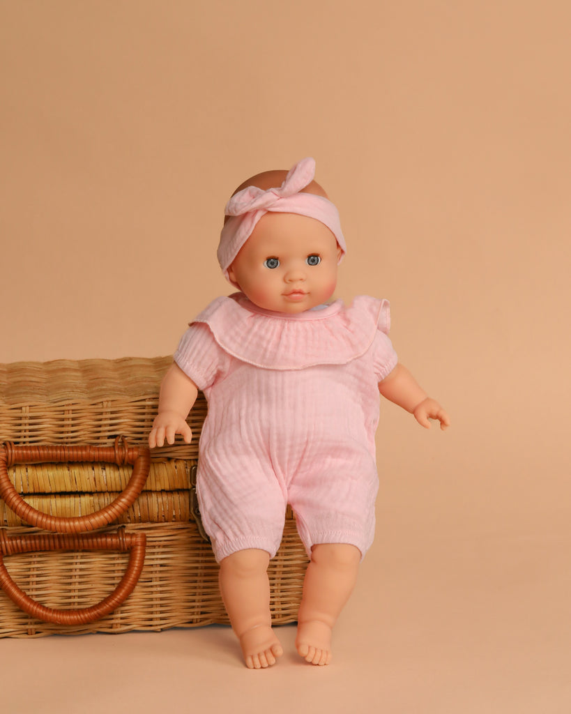 The Minikane Cuties Soft Body Doll (14") - Emilie with blue eyes, dressed in a pink romper and headband, stands by a wicker basket—perfect for pairing with your favorite doll accessories.