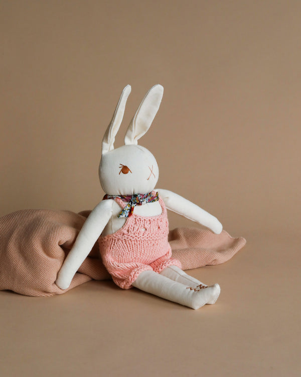 A Polka Dot Club Cream Rabbit in Hand Knit Overalls with white and peach colors, wearing baby alpaca overalls in pink and a floral neck scarf, seated against a plain beige background.