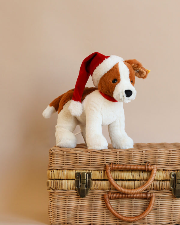 A Steiff Snuffy Puppy Dog plush, featuring a festive Santa hat, stands proudly on a woven wicker basket. The soft beige background enhances the warm tones of this charming scene.