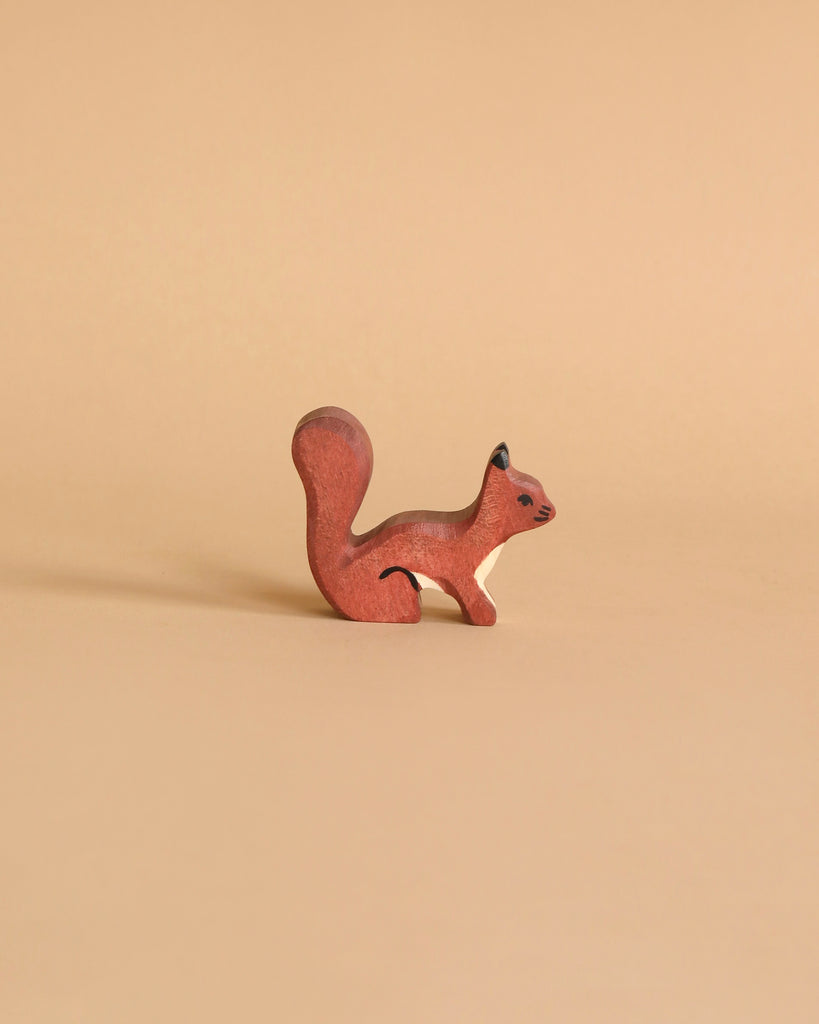 A small, handcrafted wood toy figurine of a squirrel with a bushy tail is placed on a light brown background. Made in Europe, the squirrel is painted in earthy tones of reddish-brown with minimal detailing for facial features and the texture of its fur. This charming piece is reminiscent of the Holztiger Squirrel, Standing.