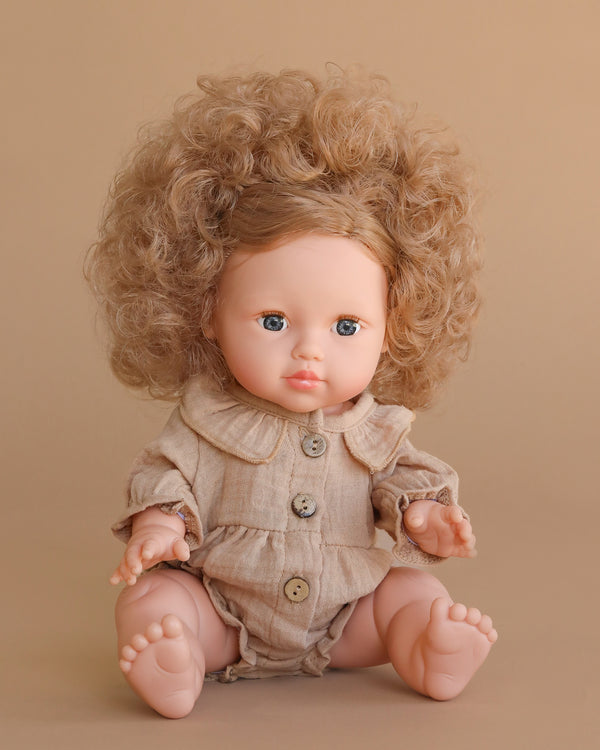The Minikane Baby Doll (13") - Lola with Clothing features curly hair, blue eyes, and a beige outfit with large buttons. This anatomically correct doll is beautifully crafted in Spain and sits against a plain beige background.