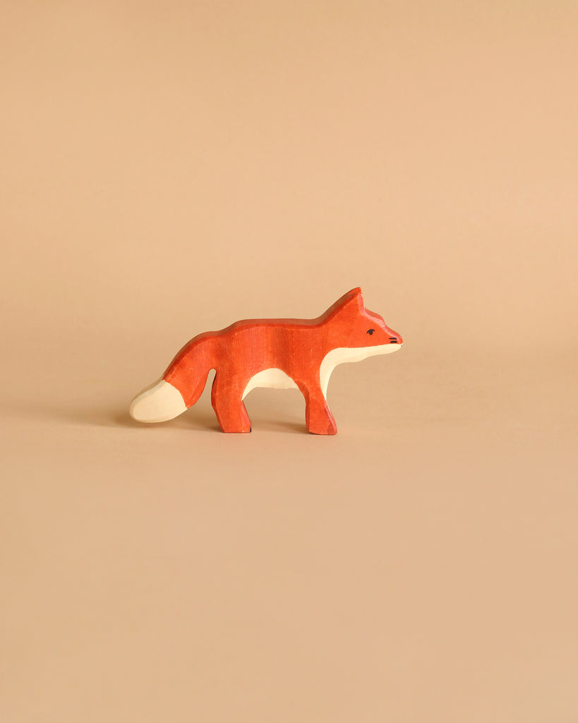 A small, handcrafted fox from the Holztiger Handmade Wooden Woodland Animals Set, featuring orange, white, and red colors, stands on a beige background—ideal for open-ended play and fans of wooden animal figures.