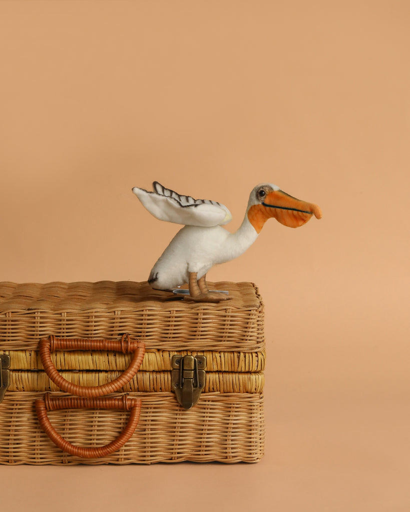 The Pelican Stuffed Animal, Small features a realistic design with an orange beak and feet, standing atop a wicker picnic basket with leather handles—one of HANSA’s beautifully hand-sewn plush animals.