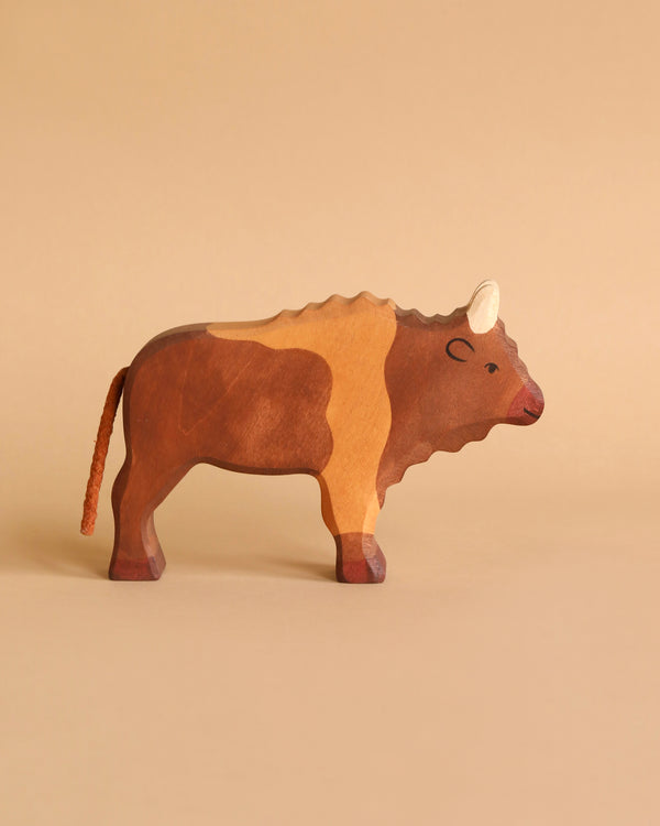 A Holztiger Bison, painted in varying shades of brown, stands against a solid beige background. Handcrafted in Europe, the bison features a detailed mane, white horns, and a tail made of twine.