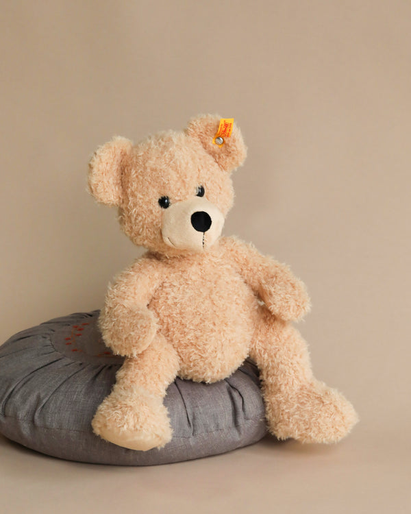A beige Steiff Fynn Teddy bear sitting on a gray cushion against a light brown background, with the Button in Ear trademark visible on its ear.