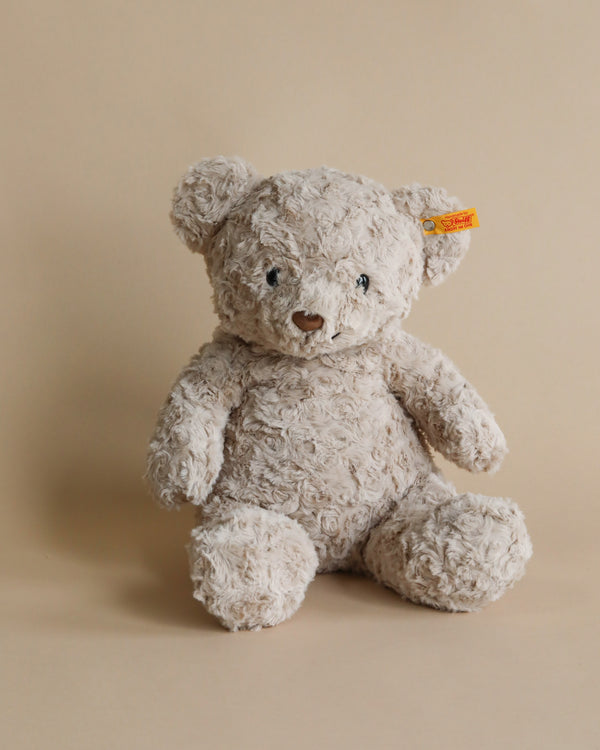 A beige, plush Steiff Honey Teddy Bear, measuring 15 inches, sits upright against a neutral background. The teddy bear features textured, curly fur and has a small yellow tag attached to its left ear. Its small dark eyes and nose give it an endearing appearance. This adorable plush toy is even machine washable for easy care.