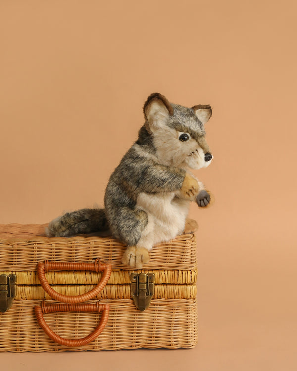 The Wolf Puppet, featuring gray and white fur from the HANSA animals collection, sits atop a woven wicker suitcase against a plain beige background.