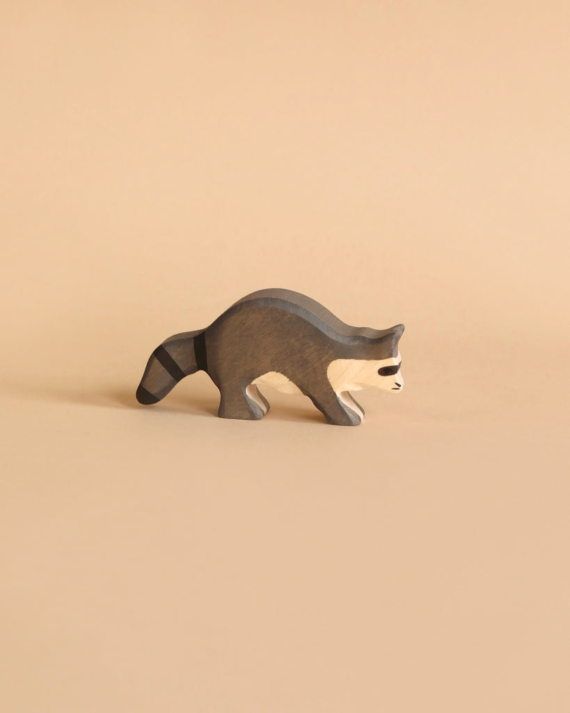 A small, minimalist wooden raccoon figurine is shown against a neutral beige background. The Holztiger Racoon is painted gray and white with characteristic black mask-like markings around its eyes and rings on its tail, showcasing the beauty of handcrafted wood. Made in Europe.