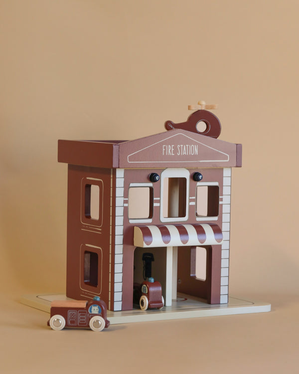 The Konges Sloejd - Wooden Fire Station, crafted from FSC-certified wood, includes a miniature fire truck and helicopter set against a beige backdrop. The station showcases a brown exterior complemented by white accents and a striped awning. Although enchanting, it contains small parts that could present a choking hazard for children under three years old.