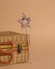 The Magic Wand - Pink Sequin Star with a pink handle leans against a closed wicker basket with leather handles and clasp, set on a plain beige background.