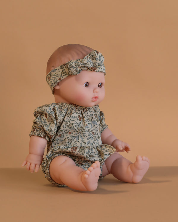 The Minikane Doll (13") - Faustine With Clothing sits on a neutral beige surface with a beige background, wearing a matching floral outfit and headband. This anatomically correct doll features short light brown hair and a realistic expression.