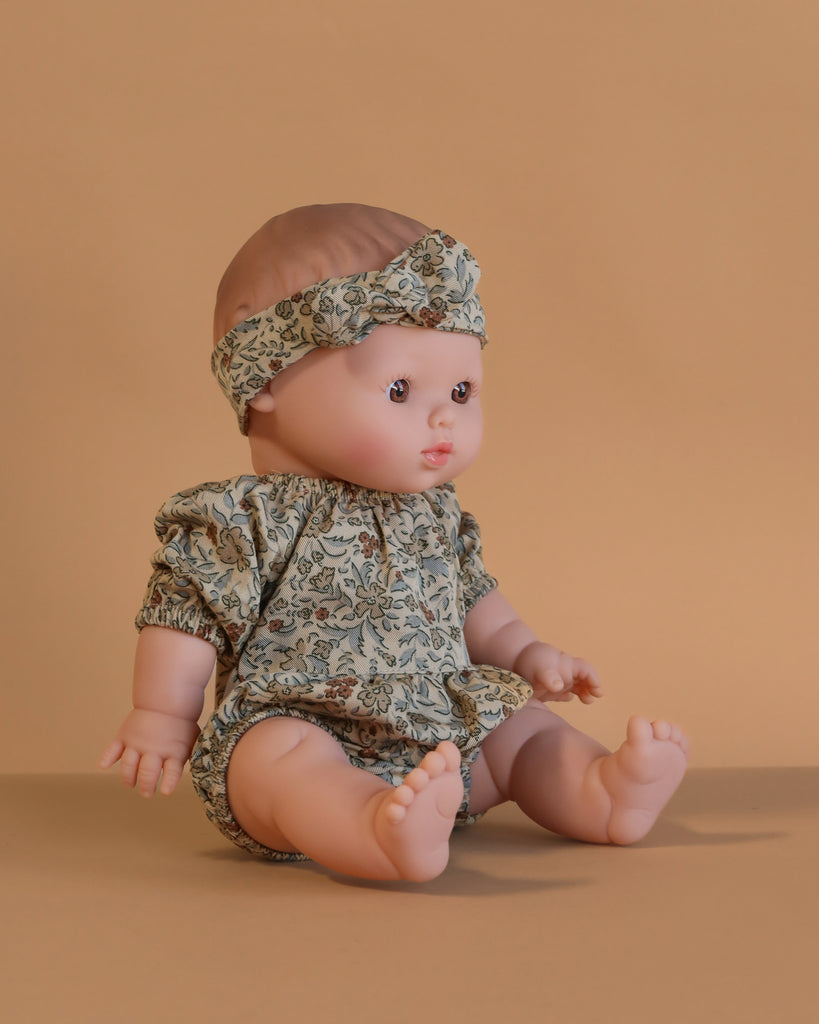 The Minikane Doll (13") - Faustine With Clothing sits on a neutral beige surface with a beige background, wearing a matching floral outfit and headband. This anatomically correct doll features short light brown hair and a realistic expression.