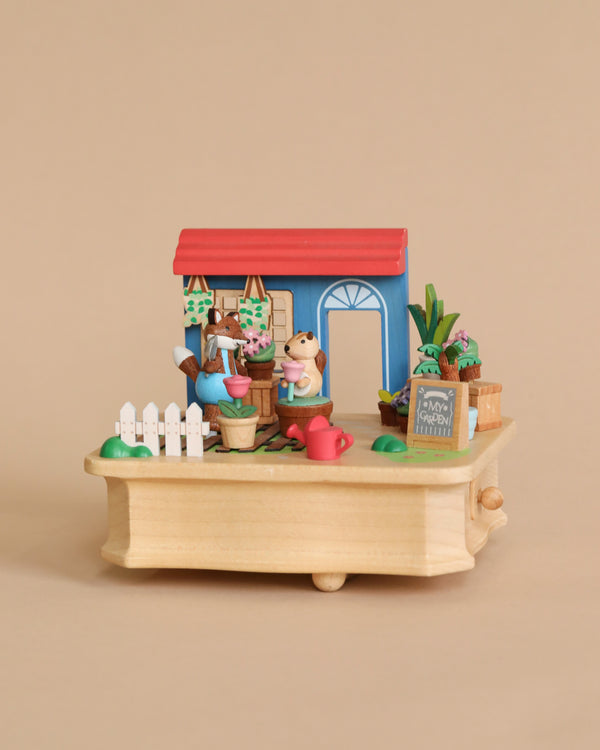 A Wooden Fox Gardener Music Box featuring a miniature scene of a toy farm stand with animal figures, including a fox and dog, surrounded by plants, vegetables, and gardening tools on a wooden platform crafted