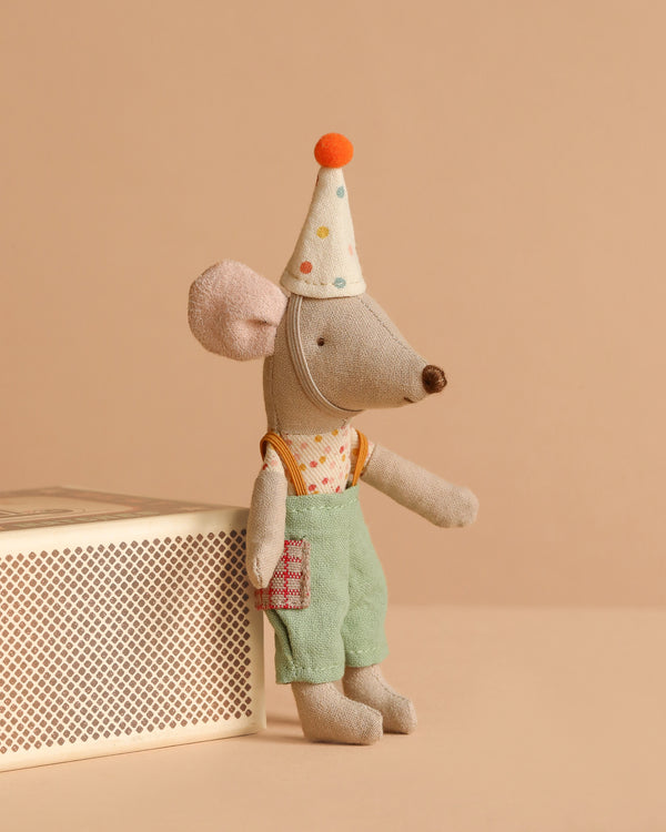 Maileg Little Sister With Clown Outfit, dressed in green overalls, a polka-dot party hat, and a printed shirt, sits on the edge of a white matchbox against a beige background.