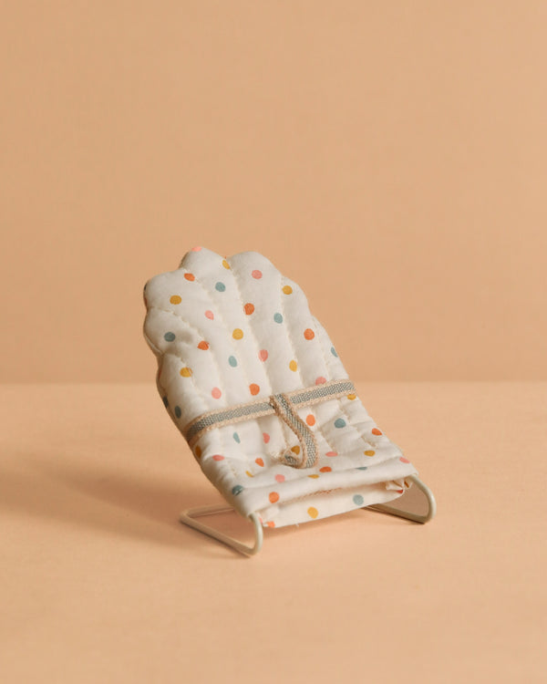The Maileg My Babysitter - Multi Dots features a small baby sitter chair with colorful polka dots on white cotton fabric and a simple metal base, set on a beige surface with a matching beige background.