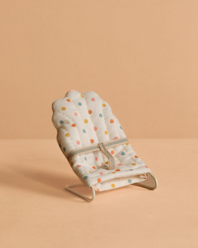 The Maileg My Babysitter - Multi Dots features a small baby sitter chair with colorful polka dots on white cotton fabric and a simple metal base, set on a beige surface with a matching beige background.