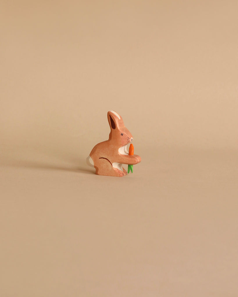 A small wooden figurine of a brown rabbit holding an orange carrot in its front paws. Handcrafted from maple and beech wood, this Holztiger Rabbit With Carrot stands out against a plain, light beige background, making the rabbit the central focus of the image. Made in Europe with exceptional craftsmanship.