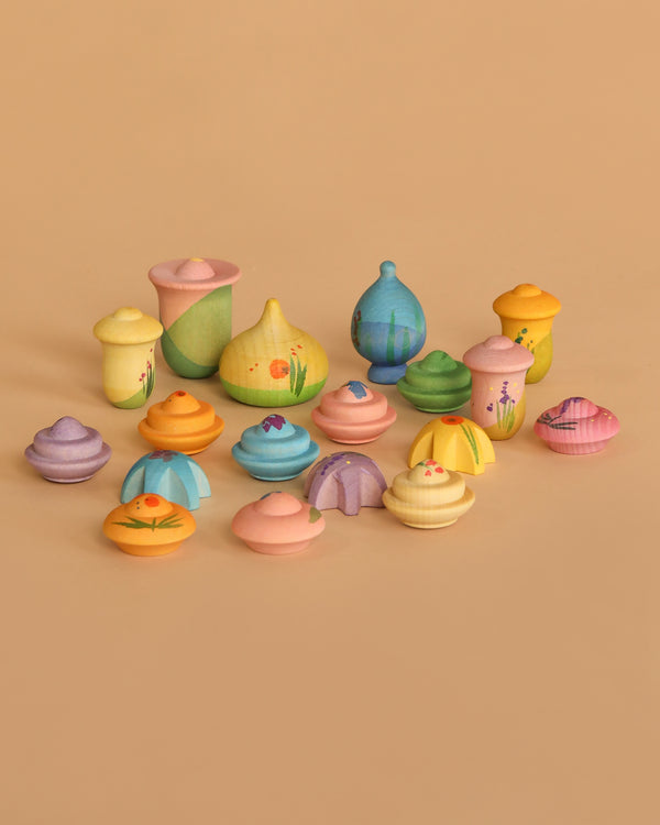 The Grapat Play Set - Flower Meadow features small, colorful, hand-painted wooden containers and lids in various pastel shapes with floral patterns, arranged on a beige surface.