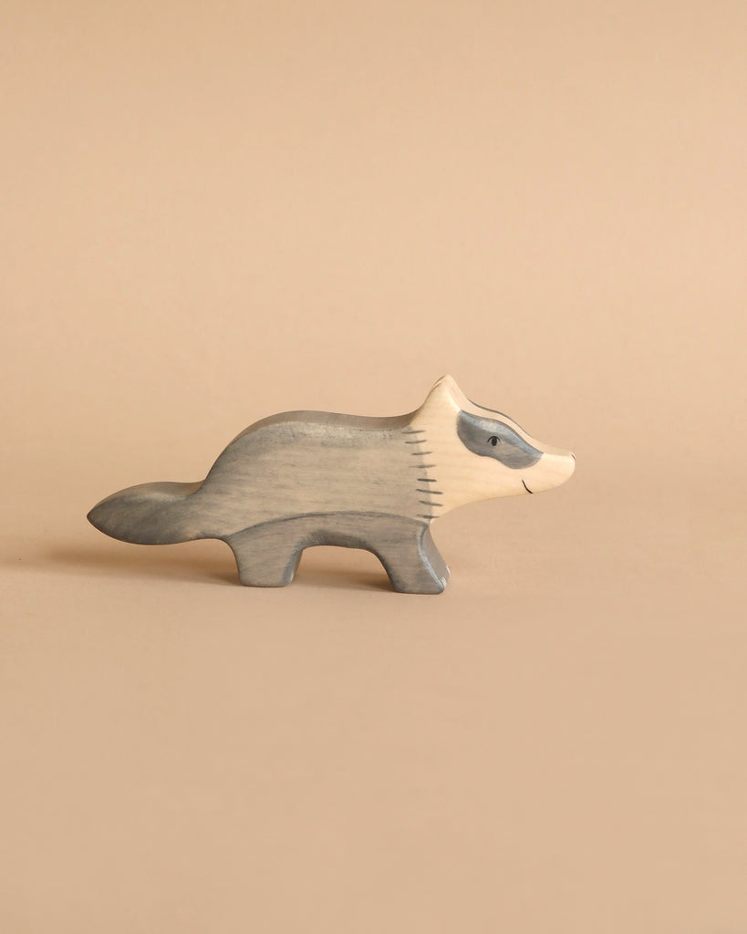 A small, handcrafted wooden figurine of a badger with a minimalist design. Made from maple and beech wood, the badger stands on all fours and is painted in shades of grey and white against a beige background. The carving style is smooth, similar to HOLZTIGER figures, giving it a simple and charming appearance.