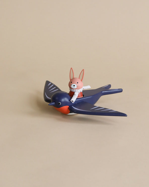 A toy rabbit named Spud the pilot Hare with red ears riding on the back of a toy blue bird named Swifty Bird against a plain beige background. The scene is playful and whimsical.