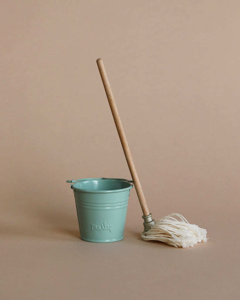 A Maileg Miniature Mop & Bucket with the word "mop" printed on it, accompanied by a bucket and mop with a wooden handle and white strands, set against a plain beige background.