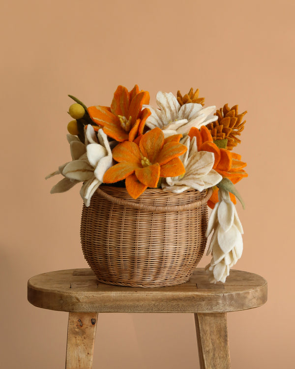 The Fall Flower Basket, featuring an everlasting wool felt bouquet of orange and white flowers in a wicker basket, is displayed on a rustic wooden stool against a plain beige background.