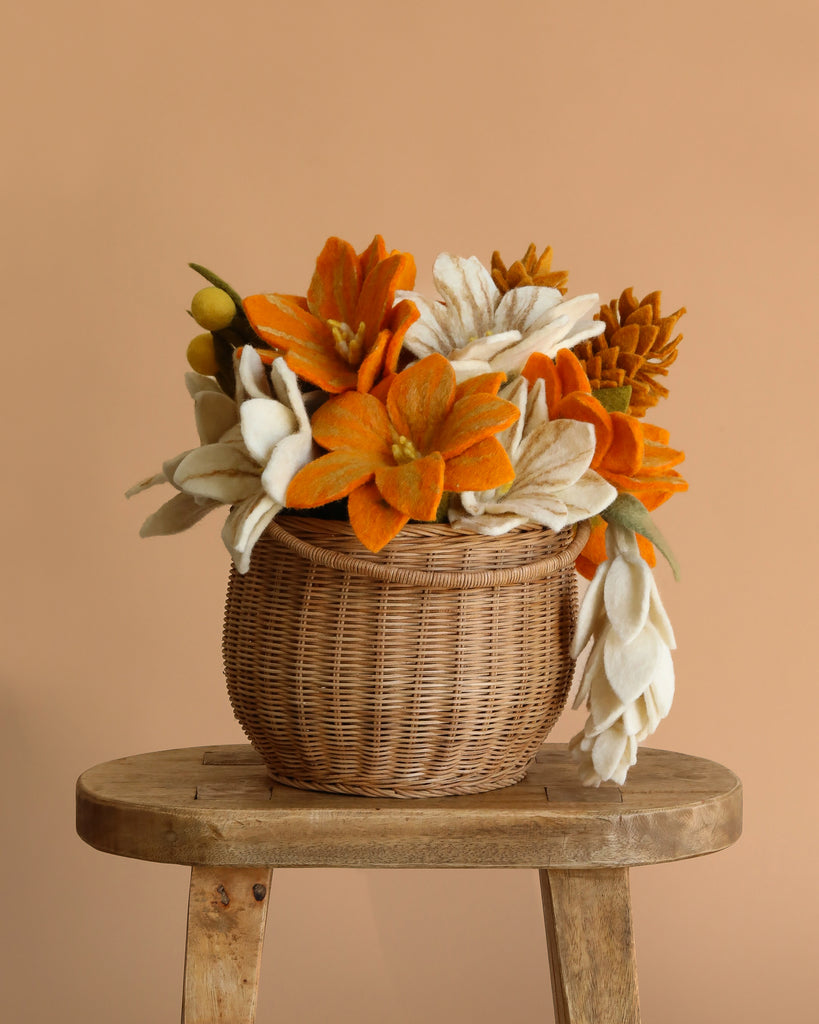 The Fall Flower Basket, featuring an everlasting wool felt bouquet of orange and white flowers in a wicker basket, is displayed on a rustic wooden stool against a plain beige background.