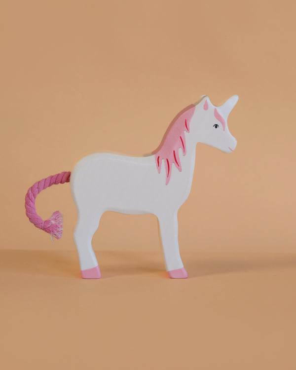 The Holztiger Unicorn is a handcrafted wooden toy with a white body, pink mane, hooves, and yarn tail. Made in Europe with exceptional detail, it stands on a peach background.
