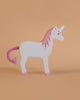 The Holztiger Unicorn is a handcrafted wooden toy with a white body, pink mane, hooves, and yarn tail. Made in Europe with exceptional detail, it stands on a peach background.