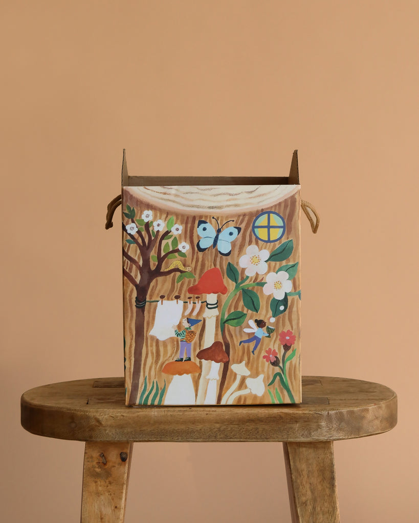 The Odin Parker x Gemma Koomen Gift Bag—a cardboard box decorated with flowers, mushrooms, a tree, butterfly, window, and hanging clothes—rests on a wooden stool against a peach background; an ideal eco-friendly gift bag.