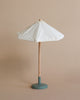 A Maileg Miniature Sunshade with a white fabric canopy and a wooden pole stands on a light blue circular base, creating a cozy atmosphere reminiscent of a terrace. The background is a neutral beige color.