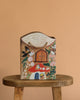The Odin Parker x Gemma Koomen Gift Bag, featuring a whimsical house shape and hand-painted florals, birds, and doors, rests on a round wooden stool against a plain beige background.