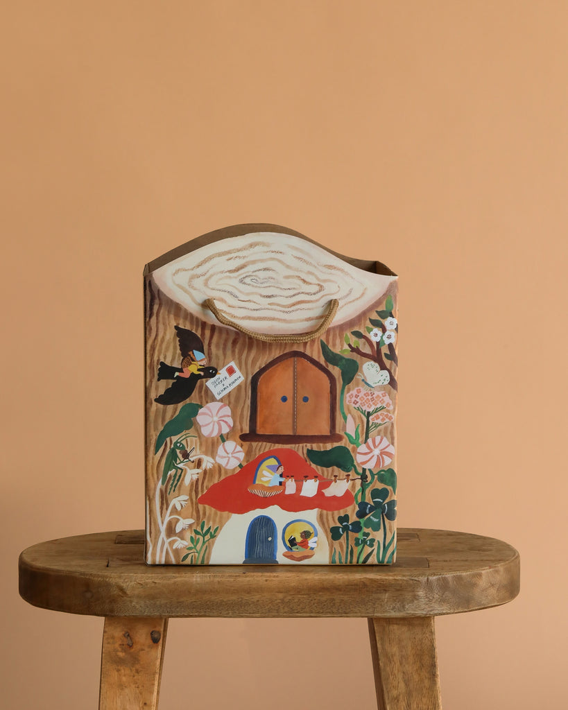 The Odin Parker x Gemma Koomen Gift Bag, featuring a whimsical house shape and hand-painted florals, birds, and doors, rests on a round wooden stool against a plain beige background.