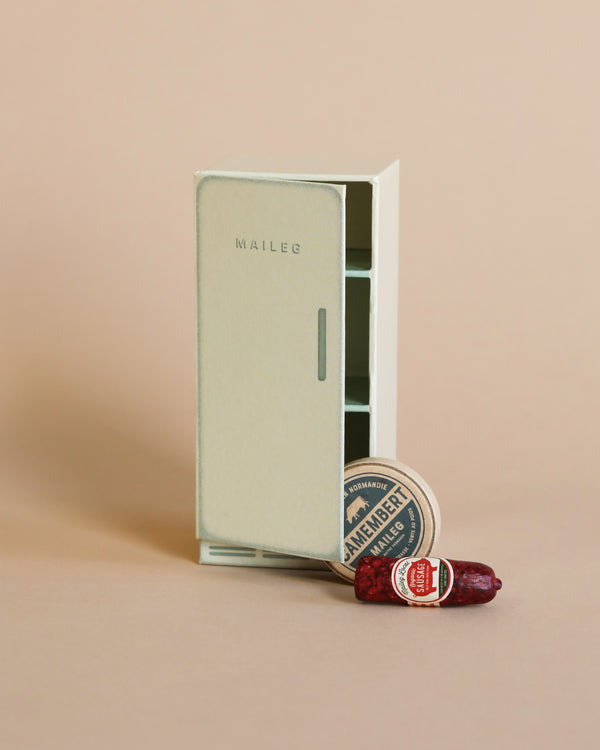 A small, elegant Maileg Cooler miniature wardrobe in off-white, displayed next to a tiny fabric sausage and a miniature can of brunswickers sardines, all set against a soft pink cardboard.