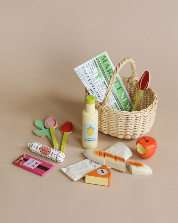 A Shopping Basket (ships in approximately one week) filled with play market items, including wooden red and yellow tulips and a rolled paper titled "Market News." Surrounding the basket are children's shopping toys such as a yellow bottle, an apple, a loaf of bread, a packet of meat, and a cheese block—perfect for pretend play.