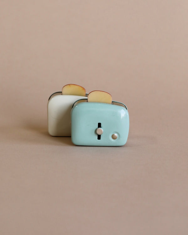 Two Maileg | Miniature Toasters, one white and one turquoise, with slices of ceramic toasted bread peeking out, set against a soft beige background.