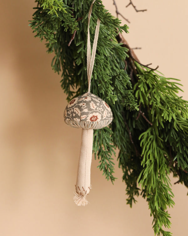 The Maileg Mushroom Ornament - Winter Flower Green, a delightful decorative fabric piece adorned with floral patterns, hangs gracefully from a green pine branch against a beige background.