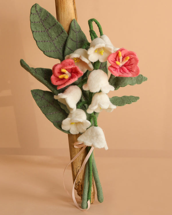The Handcrafted Felt Flower Bouquet features green leaves, white and pink felt flowers with yellow centers, tied with a pink ribbon and displayed against a wooden stick on a beige background.