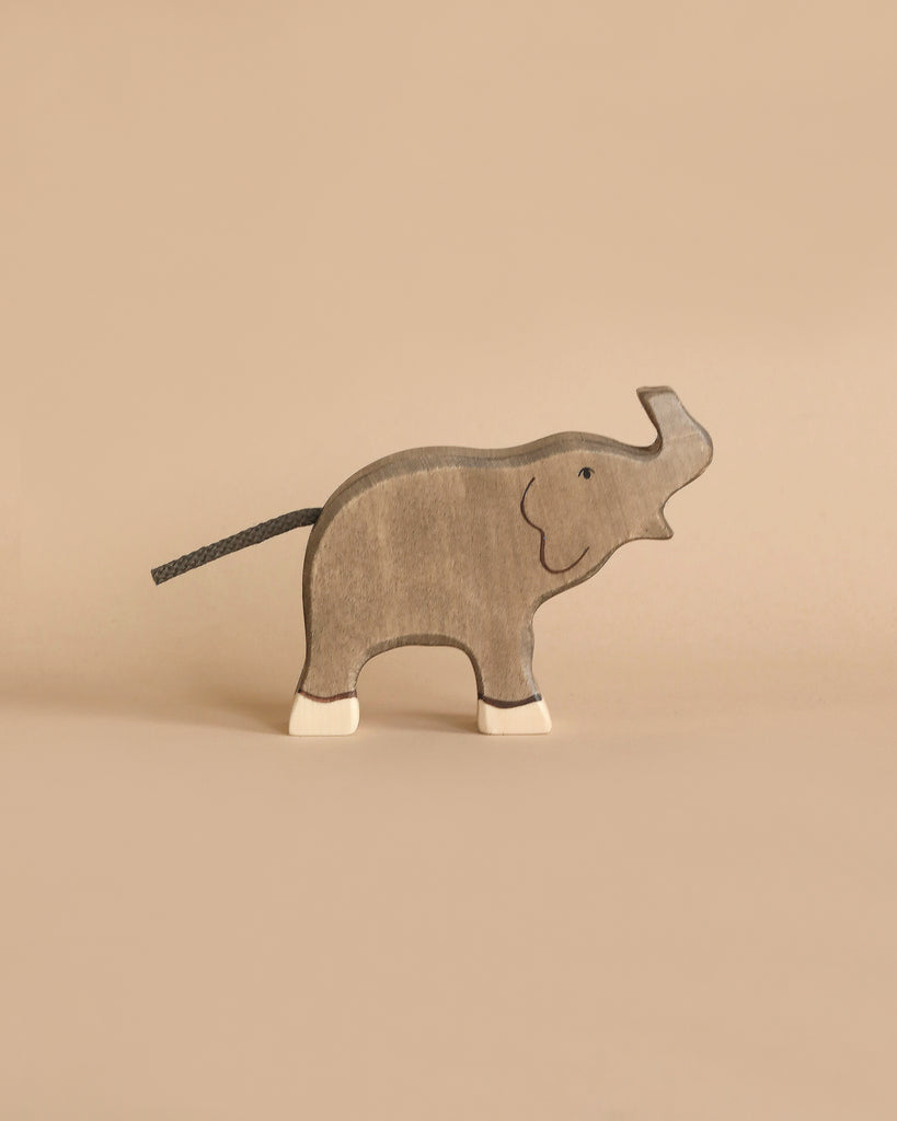 A gray, handmade wooden elephant from the Holztiger Handmade Wooden Safari Animals Set stands on a beige surface with white feet and a black tail, inspired by classic Holztiger figures and crafted from maple and beech wood.