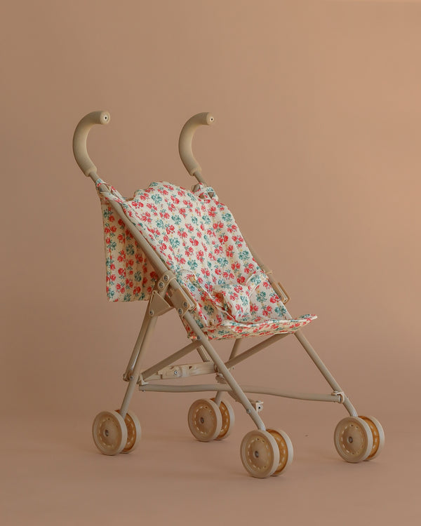The Konges Sloejd Doll Stroller - Lumina, shown against a light brown background, features a beige frame, floral-patterned removable fabric seat, four sets of double wheels, and curved handles.
