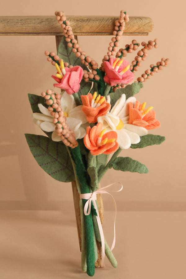 The Handcrafted Felt Flower Bouquet features pink, orange, and white blossoms with green leaves and brown berry stems, tied with a pale pink ribbon and displayed against a wooden frame on a beige background.